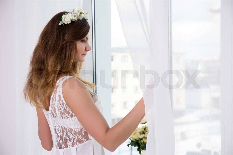 Young woman in white bridal dress standing near the window, stock photo