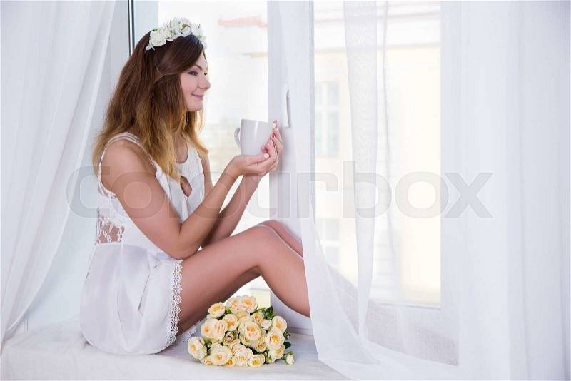 Portrait of young beautiful woman in short white dress with flowers and cup of tea sitting on window sill, stock photo