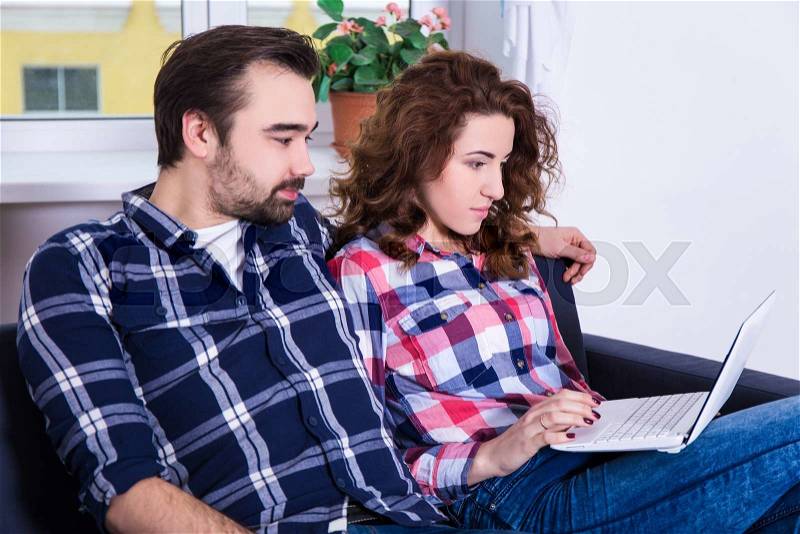 Cheerful couple searching something on laptop sitting on sofa at home, stock photo