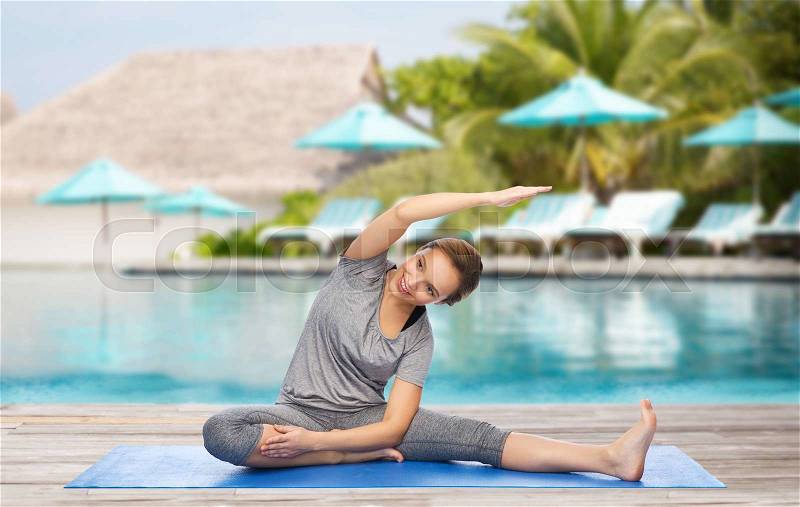 Fitness, sport, people and healthy lifestyle concept - happy woman making yoga and stretching on mat over beach and swimming pool background, stock photo