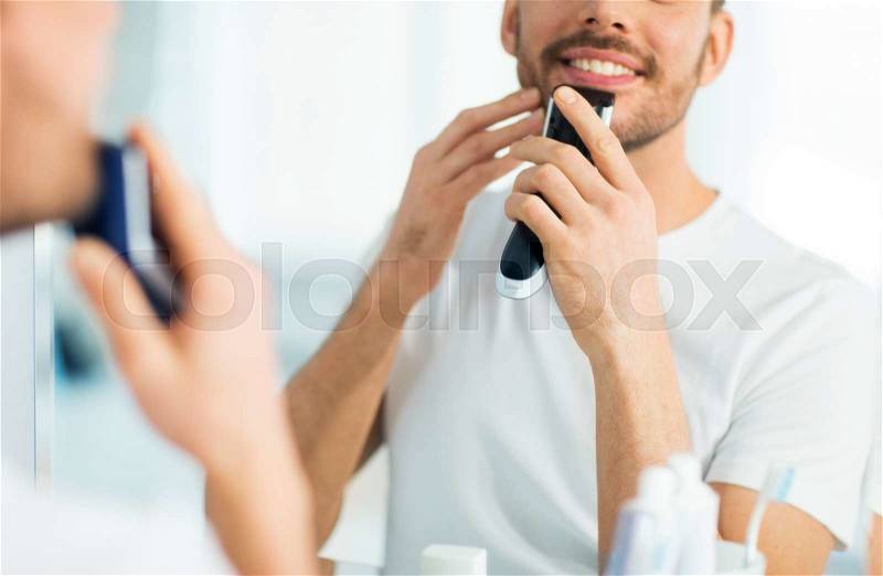 Beauty, shaving, grooming and people concept - close up of young man looking to mirror and shaving beard with trimmer or electric shaver at home bathroom, stock photo