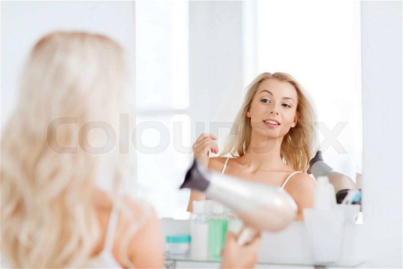 Beauty, hairstyle, morning and people concept - smiling young woman with fan blow drying her hair looking to mirror at home bathroom, stock photo