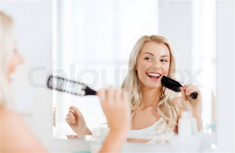 Beauty, grooming, hair care and people concept - smiling young woman looking to mirror and singing to hair brush or comb at home bathroom, stock photo