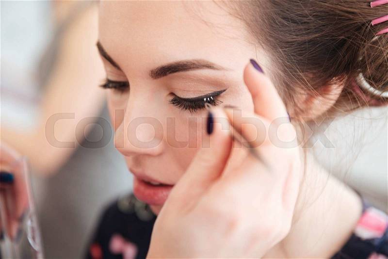 Closeup of makeup artist doing false lashes to young woman in curlers, stock photo