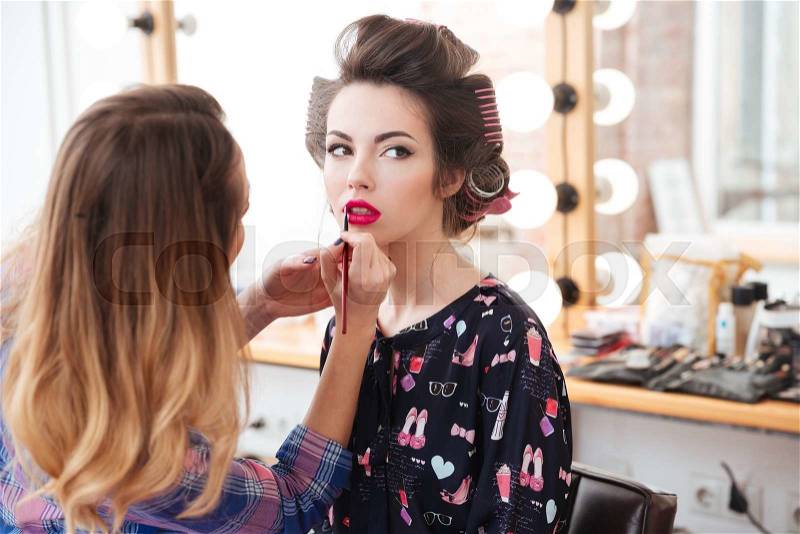 Woman makeup artist doing professional makeup with bright pink lipstick to young sensual model with curlers, stock photo