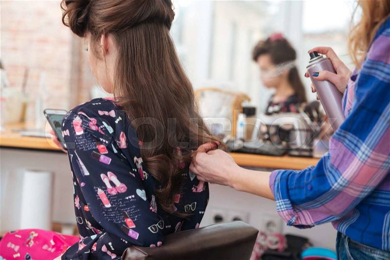 Woman hair stylist applying hairspray to long hair of young female with cell phone in beauty salon, stock photo