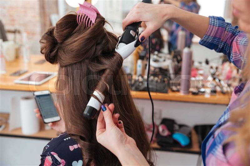Woman hairdresser making hairstyle using curling iron for long hair of young female with smartphone in beauty salon, stock photo