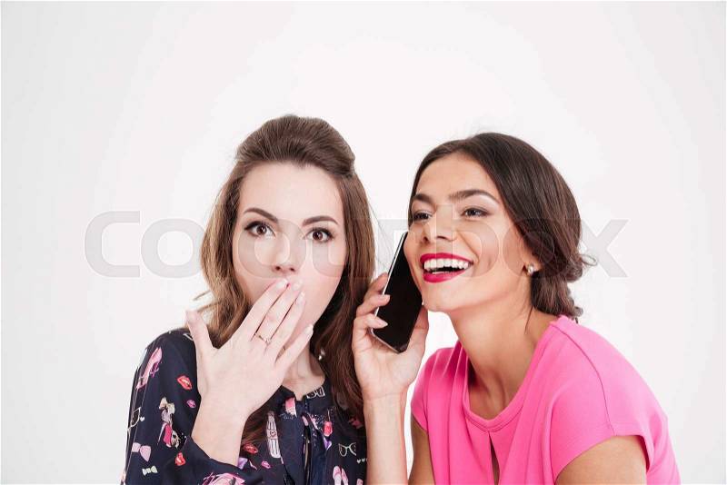 Shocked beautiful young woman overhearing conversation of cheerful female with mobile phone over white background, stock photo