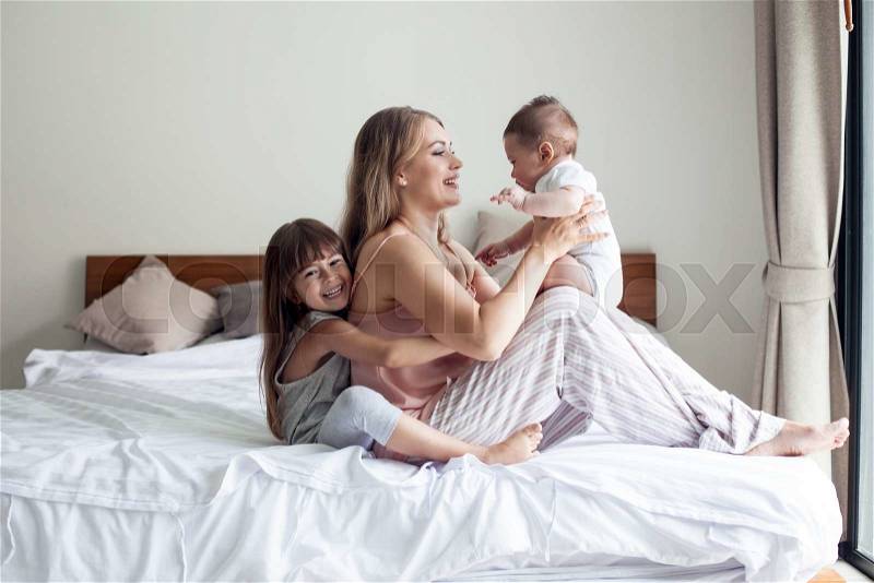 Young mom with her 5 years old dauhter and 4 months old baby dressed in pajamas are relaxing and playing in the bed at the weekend together, lazy morning, stock photo