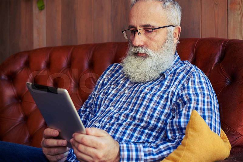 Bearded man sitting on sofa and reading news on tablet pc at home, stock photo