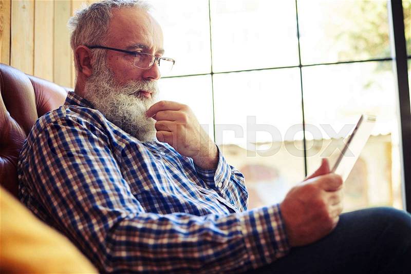 Senior man resting on sofa and reading news on tablet pc at home, stock photo