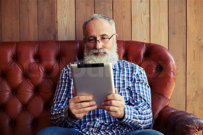 Smiley senior man sitting on sofa and reading news on his tablet pc at home, stock photo
