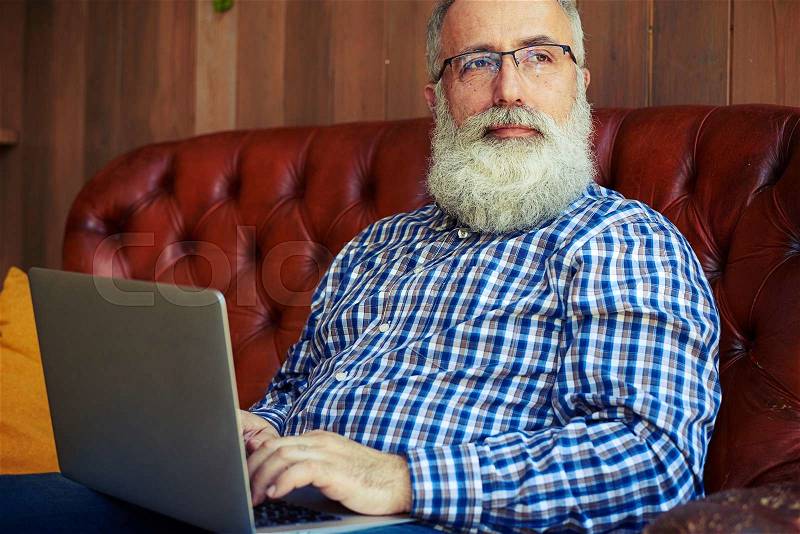 Thoughtful old man sitting with laptop and looking at window, stock photo
