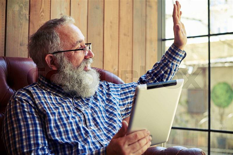 Senior man with tablet pc waving his hand and looking at the window, stock photo