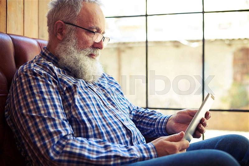 Old man resting on sofa and using tablet pc at home, stock photo