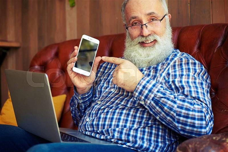 Smiley senior man pointing at smartphone and looking at camera, stock photo