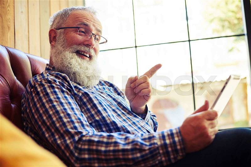 Smiley senior man sitting on the couch and pointing at tablet pc, stock photo