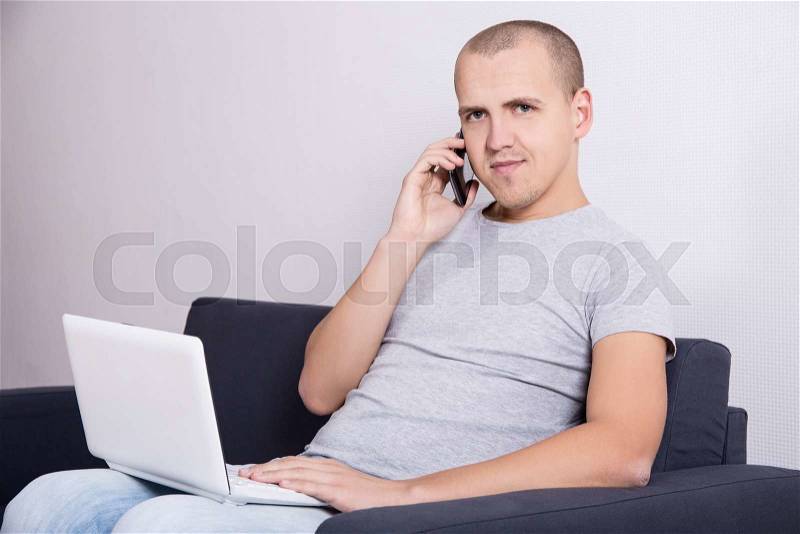 Young handsome man sitting on sofa with computer and mobile phone at home, stock photo