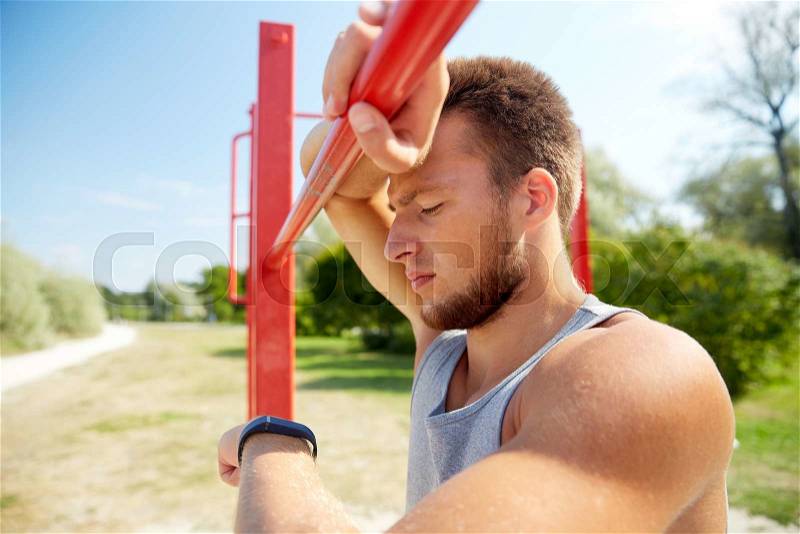 Fitness, sport, training and lifestyle concept - young man looking at heart-rate watch bracelet and exercising on horizontal bar outdoors, stock photo