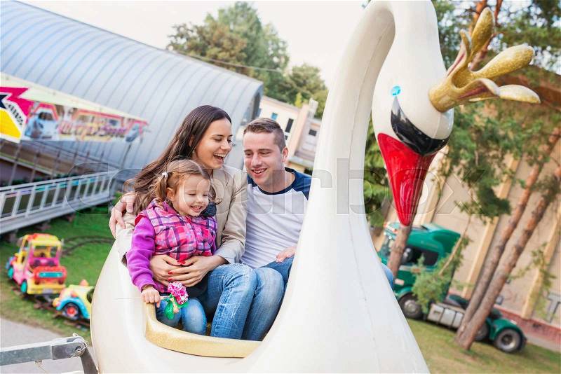 Cute little girl with her mother and father enjoying ride at fun fair, amusement park, stock photo