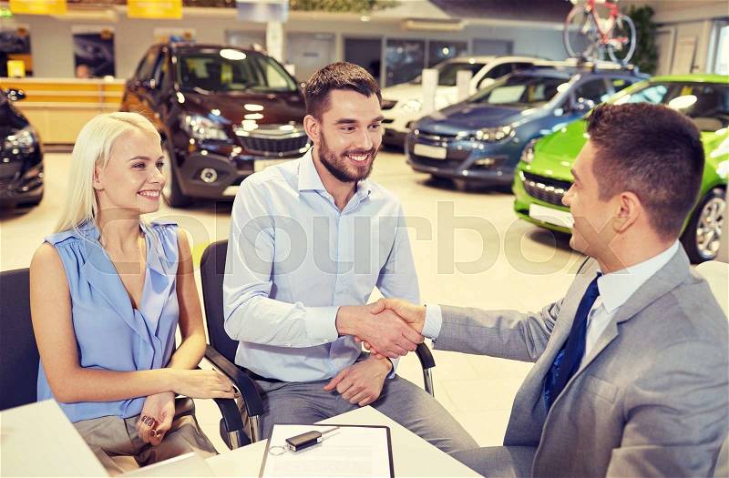 Auto business, car sale, and people concept - happy couple with dealer shaking hands in auto show or salon, stock photo