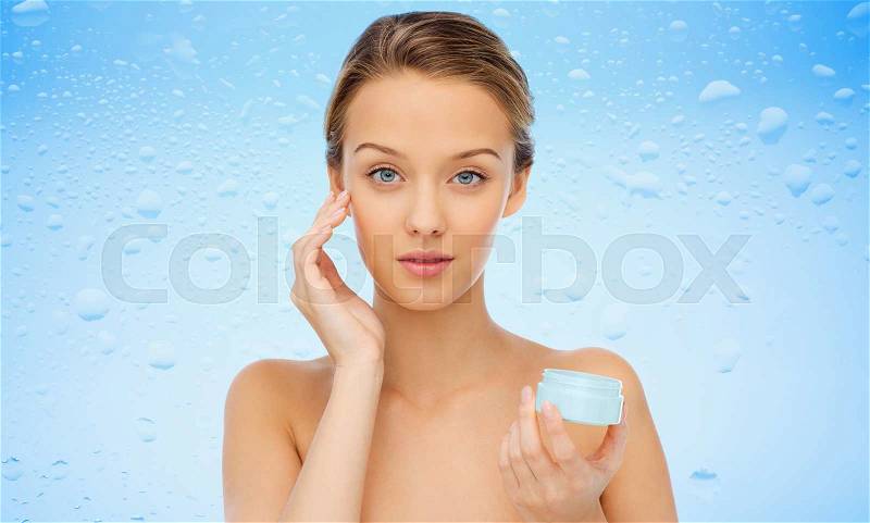 Beauty, people, cosmetics, skincare and cosmetics concept - young woman applying cream to her face over water drops on blue background, stock photo