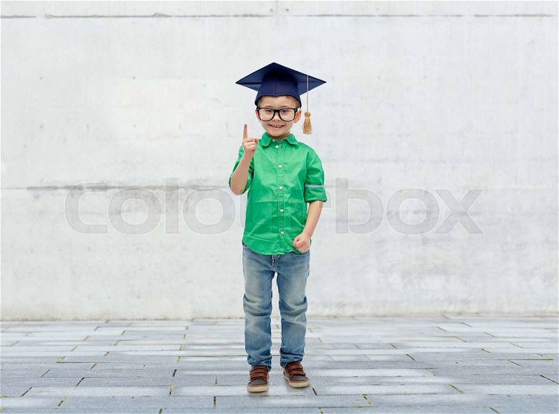 Childhood, school, education, knowledge and people concept - happy boy in bachelor hat or mortarboard and eyeglasses pointing finger up over urban concrete background, stock photo