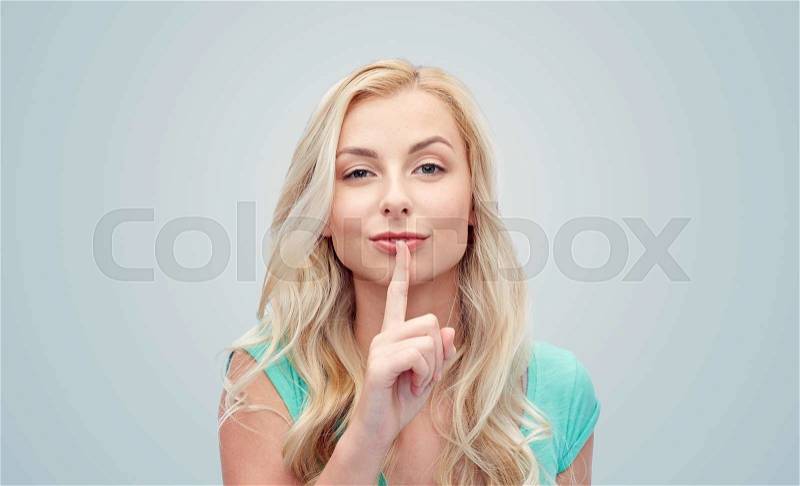 People, gesture and secret concept - beautiful young woman holding finger at her lips over gray background, stock photo