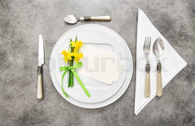 Spring table decoration with narcissus flowers. White plates, fork, knife on grey stone plate, stock photo