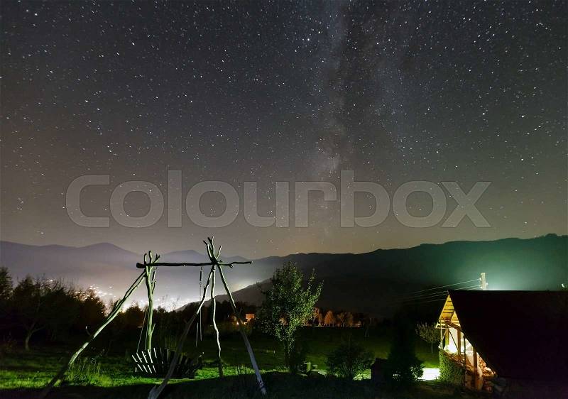Milky Way in starry night sky and rural yard illuminated in green color on mountain hill, stock photo