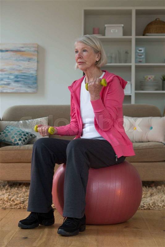 Senior lady sitting on a gym ball in her home whilst using dumbbells, stock photo