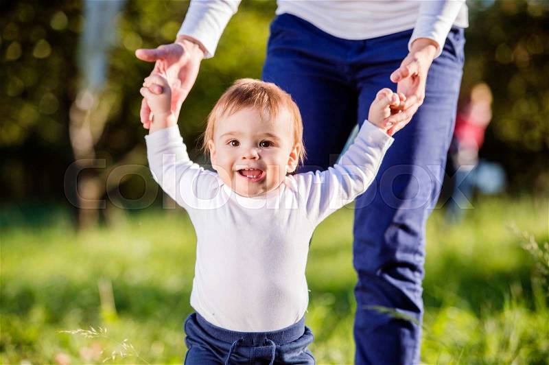 Unrecognizable mother holding hands of her little son making first steps outside in green sunny nature, stock photo