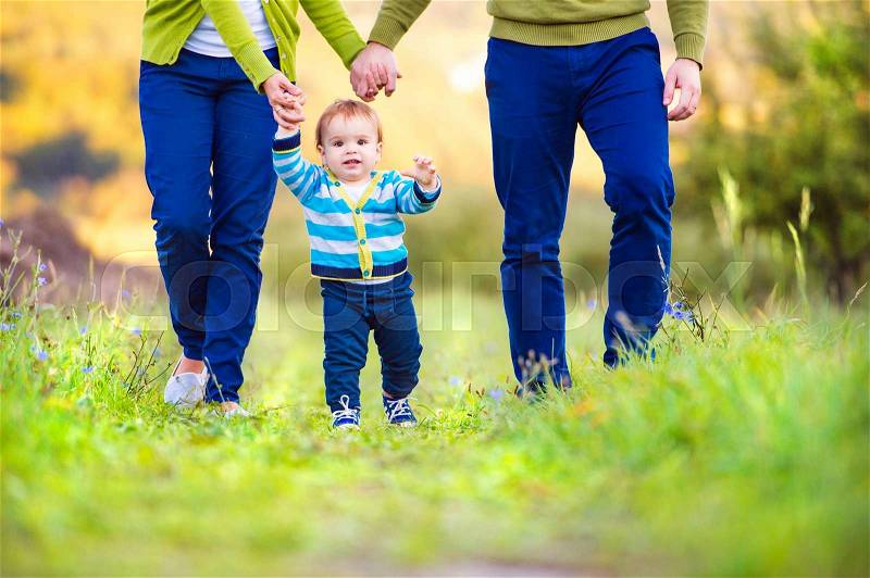 Unrecognizable mother and father holding hands of their little son making first steps outside in green sunny nature, stock photo