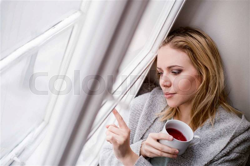 Beautiful blond woman sitting on window sill holding a cup of tea, writing on glass with finger, stock photo