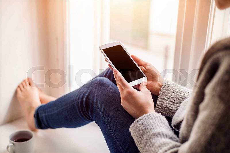 Unrecognizable woman sitting on window sill with smart phone, texting, stock photo
