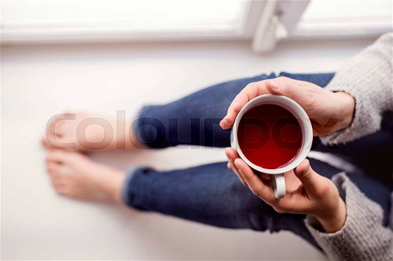 Unrecognizable woman sitting on window sill holding a cup of tea, high angle view, stock photo