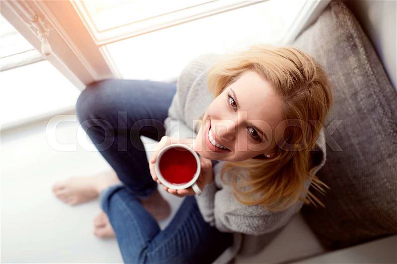 Beautiful blond woman sitting on window sill holding a cup of tea, high angle view, stock photo