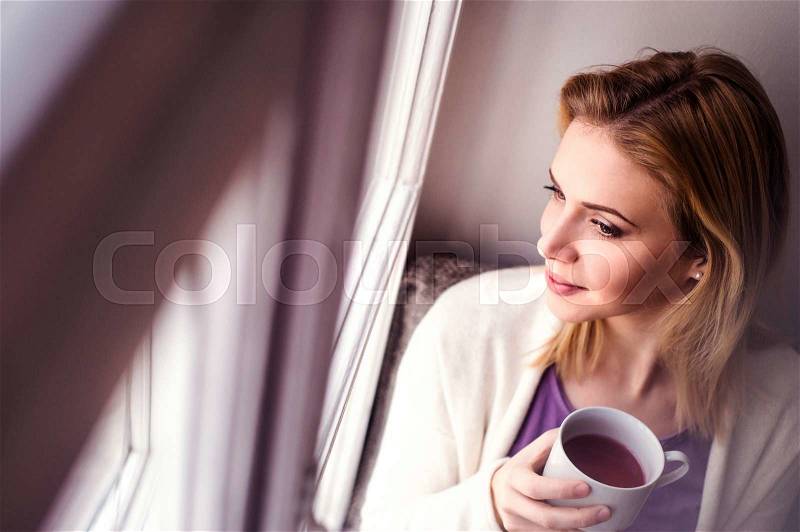 Beautiful blond woman sitting on window sill holding a cup of tea, looking out of window, stock photo