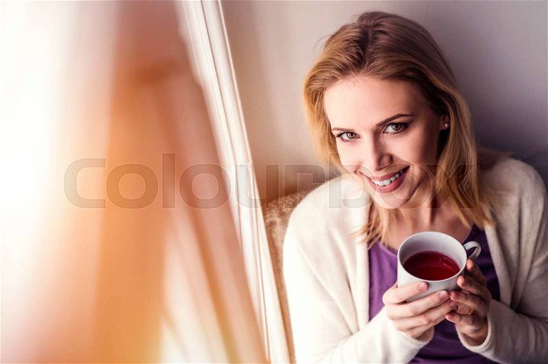 Beautiful blond woman sitting on window sill holding a cup of tea, stock photo