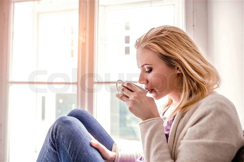 Beautiful blond woman sitting on window sill holding a cup of tea, drinking, stock photo