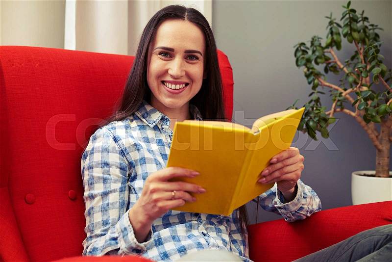 Smiley young woman resting on the chair with book and looking at camera, stock photo