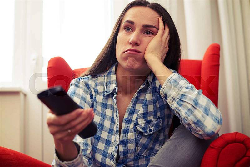 Bored young woman holding remote control and watching tv at home, stock photo