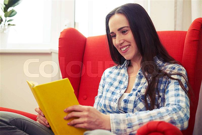 Cheerful young woman resting with book on the red chair at home, stock photo