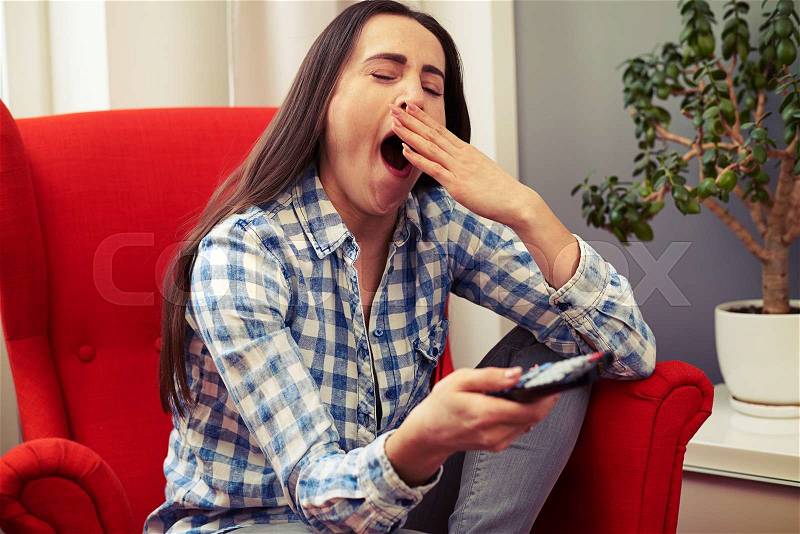 Yawning young woman sitting on the red chair and holding tv remote control, stock photo
