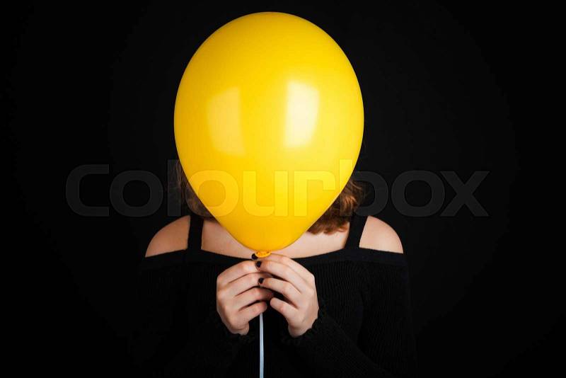 Girl hiding her face under yellow balloon, studio shot over black background, stock photo