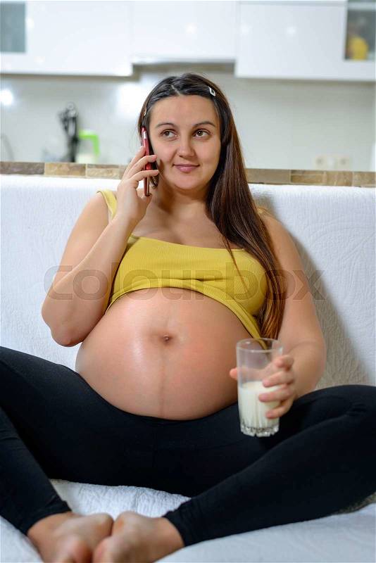 Healthy and relaxed woman in the third trimester of pregnancy sitting on a couch at home while talking on mobile phone and drinking a glass of milk, stock photo