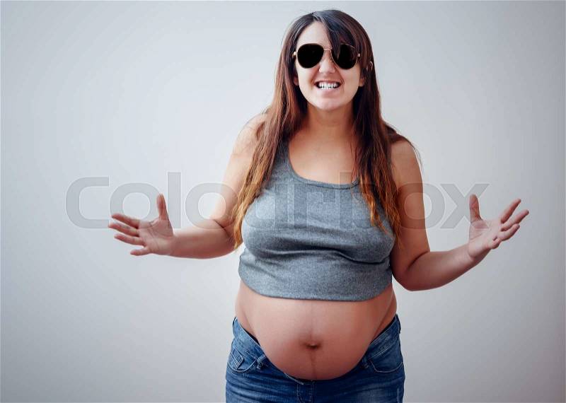 Single aggressive pregnant woman wearing gray shirt, blue jeans and sunglasses while gesturing with hands over gray background and copy space, stock photo