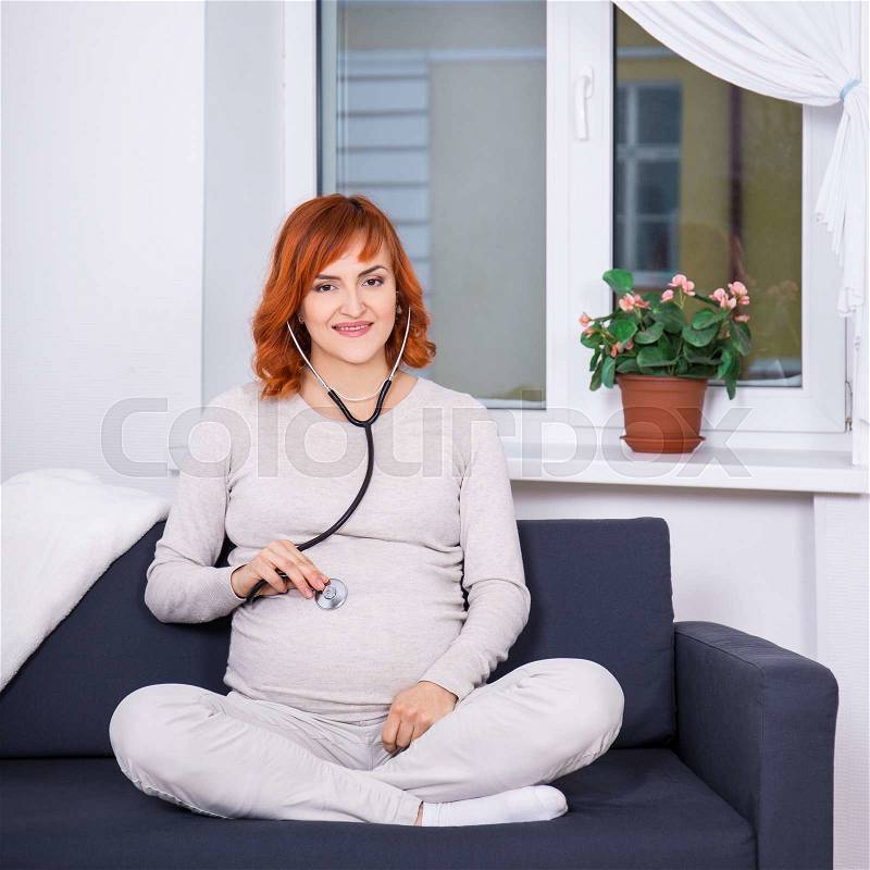 Young pregnant woman listening baby\'s heartbeat with stethoscope at home, stock photo
