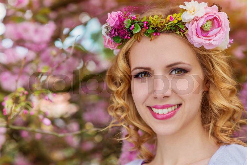 Smiling woman with blond hair with flower wreath against pink tree in blossoom, spring nature, stock photo