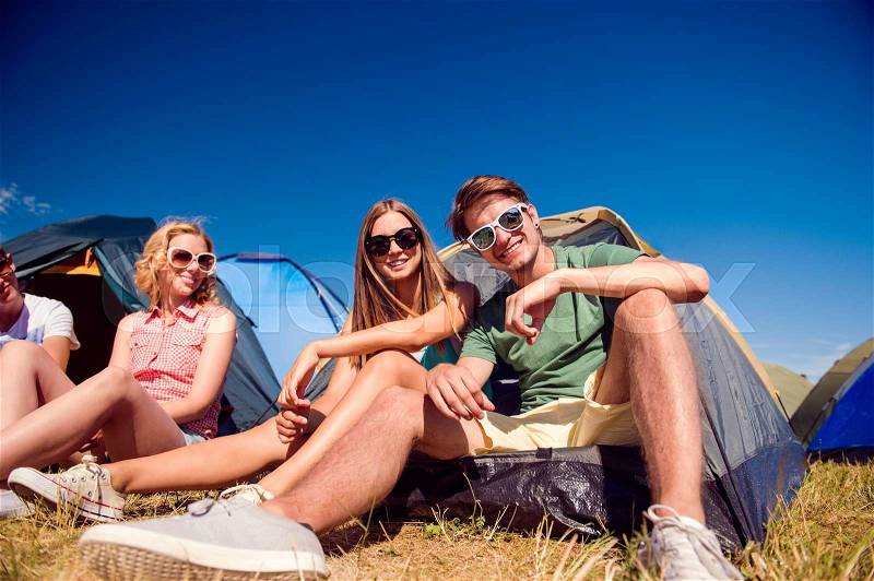 Group of teenage boys and girls at summer music festival, sitting on the ground in front of tents, stock photo